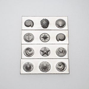 May include: A collection of silver-toned rectangular decorative tiles, each featuring a different nautical-themed design. The designs include seashells, an anchor, a compass rose, a starfish, a crab, a turtle, and swirling patterns. The tiles are arranged in a grid.