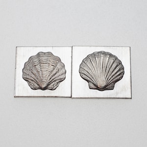 May include: Two silver-colored metal molds, each with a seashell design. One mold features a scallop shell, and the other has a fan-shaped shell. The molds are rectangular and have a slightly distressed finish.
