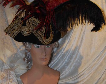 May include: A black tricorn hat with gold trim and red and black feathers. The hat is worn on a mannequin with a white wig.