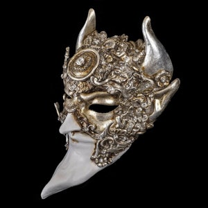 Puede incluir: Una máscara de carnaval plateada y dorada con una nariz larga y puntiaguda y detalles ornamentados. La máscara tiene una cara pintada de blanco y presenta un par de cuernos.