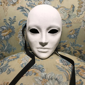 Può includere: Una maschera bianca da carnevale con buchi neri per gli occhi e un nastro nero attaccato. La maschera è appoggiata su un tessuto con motivo floreale.