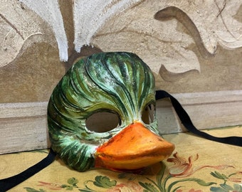 May include: A green and orange duck mask with a black ribbon. The mask is made of paper mache and has a realistic look.