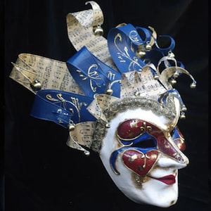 May include: A white and gold Venetian masquerade mask with blue and red accents. The mask features a jester's hat with musical notes and bells.