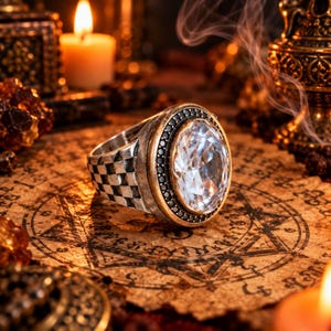 Può includere: Un anello color argento con una grande pietra preziosa trasparente dal taglio ovale incastonata in una montatura dorata. La fascia presenta un motivo a scacchiera testurizzato e l'incastonatura è circondata da piccole pietre scure.