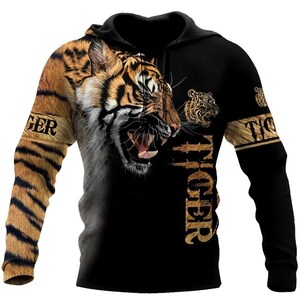 Op de afbeelding: Zwarte hoodie met een opvallend tijgerontwerp. Het gezicht van de tijger staat centraal, met oranje en zwarte strepen. Het woord "TIGER" staat op de mouwen. De hoodie heeft een capuchon en lange mouwen.
