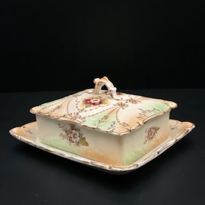 Pode incluir: Uma manteigueira de porcelana vintage com tampa e base retangular a condizer. O prato apresenta um design floral com cores pastel, incluindo rosa, amarelo e verde, e acabamento dourado. A tampa tem uma pequena pega.