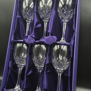 Puede incluir: Un juego de seis copas de vino de cristal transparente con intrincados patrones tallados, presentadas en una caja forrada de satén púrpura. Las copas tienen tallos largos y una forma de cuenco clásica.