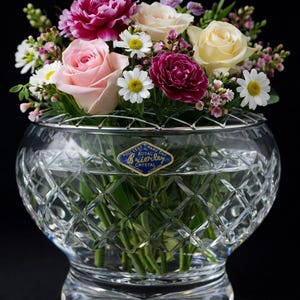Op de afbeelding: Een kristallen vaas met een ruitpatroon, gevuld met een kleurrijk bloemstuk. Het boeket bevat roze, witte en gele rozen, madeliefjes en andere bloemen. Een blauw-wit etiket is zichtbaar.