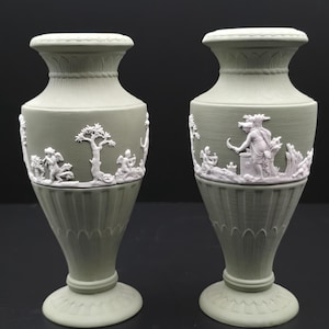 Może przedstawiać: Dwa szałwiowe zielone wazony ceramiczne z białymi reliefami. Wazony przedstawiają klasyczne sceny z postaciami, drzewami i elementami architektonicznymi. Mają pękaty kształt z rozszerzoną krawędzią i podstawą na cokole, prezentując neoklasyczny styl.