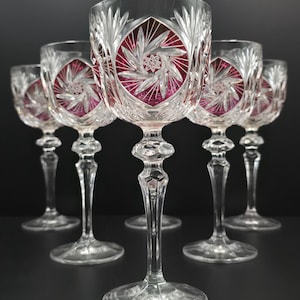 May include: A set of five clear crystal wine glasses featuring a faceted stem and a bowl decorated with a deep red circular medallion. Each medallion displays a detailed starburst pattern cut into the glass, contrasting with the clear crystal.