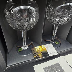 Peut inclure: Deux verres à vin en cristal clair avec des motifs gravés complexes, présentés dans une boîte noire. Les verres ont une forme de bol arrondie et une longue tige. La boîte contient une carte avec "living with Waterford Crystal".