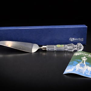 Puede incluir: Un servidor de pastel plateado con un mango de cristal transparente y una caja azul. El mango tiene un diseño facetado con un extremo decorativo. También hay una pequeña tarjeta con las palabras "Waterford Crystal".