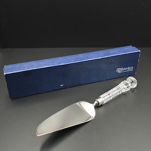 Puede incluir: Un servidor de pastel plateado con un borde dentado y un mango de cristal transparente y texturizado. El servidor está junto a una caja azul con el logotipo "Waterford".