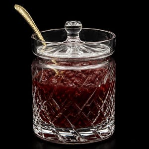 May include: A clear glass jar with a lid, filled with a dark red preserve. The jar has a diamond-cut pattern and a small gold spoon rests inside. The lid has a small knob for easy opening. The background is black.
