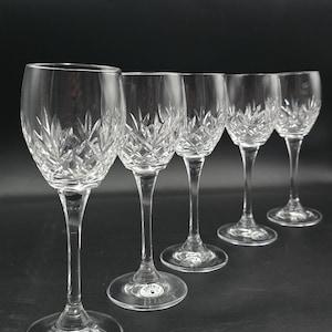 May include: Five clear crystal wine glasses with intricate cut patterns. The glasses have long stems and round bases, and are arranged in a row against a dark background. Each glass has a label that says "Bohemia".