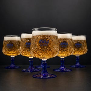 Puede incluir: Cinco copas de cerveza de cristal transparente con un patrón de diamantes texturizado en el cáliz y un tallo y base de color azul sólido. Cada copa contiene cerveza dorada con espuma blanca y el texto azul 1664 BLANC en el frente.
