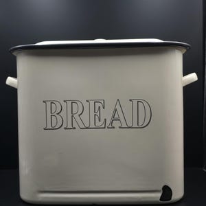 May include: A cream-colored bread box with a black rim and handles. The word "BREAD" is printed in large, black capital letters on the front. The box has a rectangular shape and a lid.