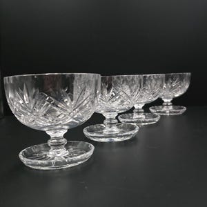 Op de afbeelding: Vier heldere kristallen dessertkommen met ingewikkelde geslepen glazen patronen. Elke kom heeft een afgeronde vorm, een korte steel en een cirkelvormige basis. De kommen zijn in een rij gerangschikt en tonen hun elegante ontwerp.