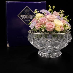 May include: A cut-glass flower vase with a silver metal grid, holding a bouquet of pink, purple, and yellow roses. The vase is on a pedestal base. A dark blue box with "Edinburgh Crystal" is in the background.