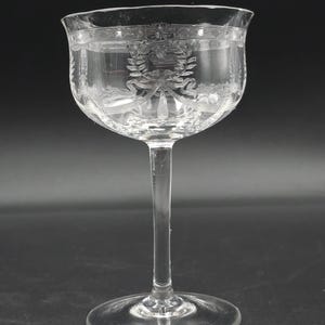 May include: A clear, stemmed champagne glass with etched floral and ribbon designs. The glass has a wide bowl and a long, slender stem. The etched details include a wreath and ribbon motif, adding a touch of elegance to the glassware.