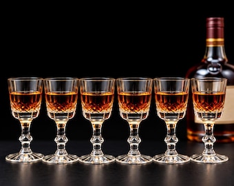 May include: Six clear glass stemmed glasses filled with amber liquid, each featuring a diamond-cut pattern on the lower bowl and a fluted base. A glass bottle with a dark red cap and amber liquid is positioned behind the glasses.
