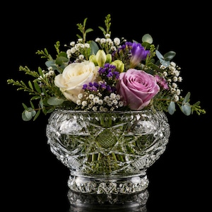 Könnte beinhalten: Eine klare, geschliffene Glasschale als Vase mit einem komplexen geometrischen Diamant- und Sternmuster, gefüllt mit einem Blumenarrangement aus weißen und lavendelfarbenen Rosen, violettem Statice, weißem Schleierkraut und Eukalyptus.