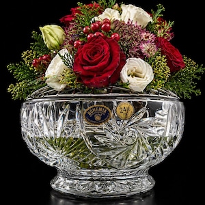 Op de afbeelding: Een heldere kristallen kom met een bloemstuk. Het arrangement bevat rode rozen, witte bloemen en groen. De kom heeft een decoratief bladerpatroon en een Bohemia-label.