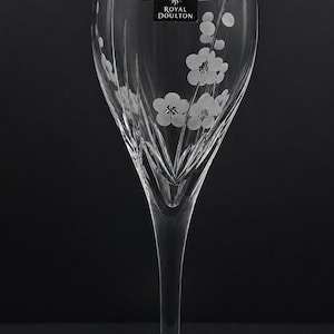 May include: A clear crystal wine glass with etched floral designs. The glass has a long stem and a wide bowl, with a delicate pattern of flowers and leaves. The Royal Doulton logo is visible on the glass.