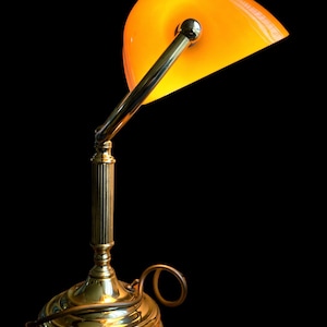 Puede incluir: Una lámpara de escritorio de latón vintage con una pantalla naranja en forma de media luna. La lámpara tiene una columna estriada y una base redonda. La pantalla está inclinada, sobre un fondo negro.