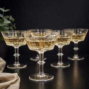 Könnte beinhalten: Sechs klare Champagner-Coupe-Gläser gefüllt mit einer funkelnden goldenen Flüssigkeit. Die Gläser haben ein detailliertes, strukturiertes Muster auf der Schale und ein klassisches Stiel-Design. Der Hintergrund ist dunkel und hebt die Gläser hervor.