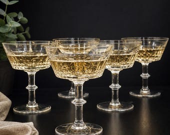 May include: Six clear champagne coupe glasses filled with a sparkling golden liquid. The glasses have a detailed, textured pattern on the bowl and a classic stem design. The background is dark, highlighting the glasses.