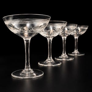 Op de afbeelding: Een set van vier heldere glazen coupes opgesteld in een rij. Elk glas heeft een brede, ondiepe kelk met een delicaat gegraveerd bloemmotief en een horizontale band van decoratieve lussen nabij de rand, rustend op een slanke steel en voet.