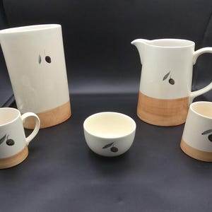 Könnte beinhalten: Ein weißes Keramik-Geschirrset mit einer hohen Vase, einem Krug, zwei Tassen und einer kleinen Schale. Jedes Stück hat einen braunen Sockel und ist mit einem schwarzen Olivenzweig-Design verziert. Das Set eignet sich zum Servieren von Getränken.