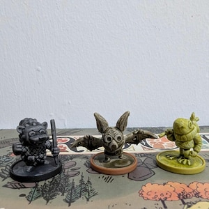 May include: Three fantasy game miniatures. A dark gray creature with a staff, a bat-like figure with outstretched wings, and a green frog-like character. Each figure is on a circular base, set on a game board.
