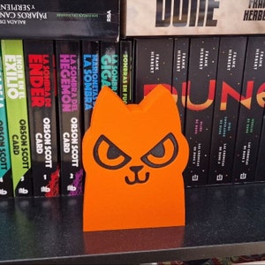 May include: Orange cat-shaped bookend with black eyes and a black nose. The bookend is sitting on a shelf with a row of books behind it. The books have titles like "Dune" and "Ender's Game".
