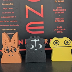 May include: Three colorful, stylized game pieces in orange, black, and yellow are displayed in front of a book cover. The orange piece resembles a rabbit, the black piece a cat, and the yellow piece a frog. The background features text in Spanish.