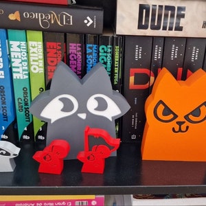 May include: Three small, colorful, 3D printed figurines of animals. A gray raccoon, a red cat, and an orange cat with angry eyes. The figurines are sitting on a shelf in front of a bookshelf filled with books.