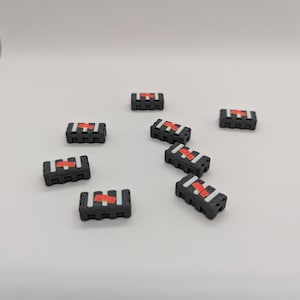 Puede incluir: Una colección de pequeños objetos rectangulares negros con una franja roja y detalles blancos. Los objetos están dispersos sobre una superficie blanca, con algunos en primer plano y otros más atrás. Parecen ser contenedores o cajas en miniatura.