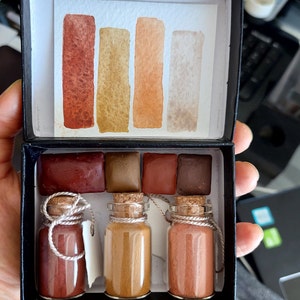 May include: A set of four watercolour paints in a black box with a white paper insert showing the colours. The colours are a deep red, a brown, an orange, and a light beige. The paints are in small glass jars with cork stoppers and twine.