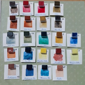 Op de afbeelding: Een verzameling van 24 aquarelverfpannetjes, gerangschikt in vier rijen van zes. De pannetjes zijn gelabeld met nummers en namen, zoals Hematiet, Ultramarijnroze, Chinacridonroze, Oker en Kobaltblauw. De pannetjes zijn gerangschikt op een groene ondergrond.