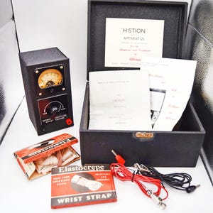 May include: A vintage Histion apparatus in a black case with a diagnostic meter, instruction manuals, and Elastocrepe wrist strap packaging. The set includes red and black electrical leads with metal clips for medical allergy treatment.
