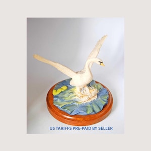 Può includere: Una statuetta di cigno bianco con le ali spiegate, posata su una base di ninfea. La base è un mix di blu, verde e giallo, su un supporto rotondo in legno. Il cigno ha un becco giallo. Il testo "US TARIFFS PRE-PAID BY SELLER" è in basso.