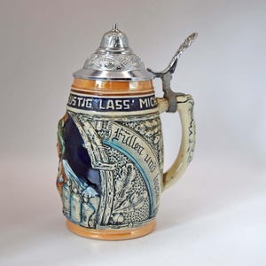 Peut inclure: Une chope à bière en céramique avec un couvercle et une anse métalliques. Le corps présente des motifs en relief, notamment des feuilles de chêne et le texte LUSTIG LASS' MICH FÜLLEN UND LEEREN. La chope arbore des tons crème, bleu et orange.