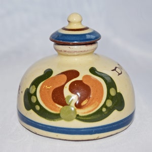 Puede incluir: Un tarro de cerámica blanca con tapa, decorado con un diseño floral marrón, naranja y verde. El tarro tiene una banda azul alrededor de la parte superior e inferior. La tapa también está decorada con una banda azul.