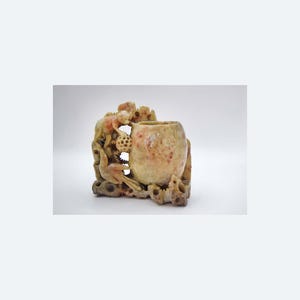 Puede incluir: Un jarrón de piedra tallada con una combinación de colores marrón claro y rosa. El jarrón presenta intrincadas tallas de un pájaro, un mono y otros elementos decorativos. Tiene una abertura redonda y una superficie texturizada.