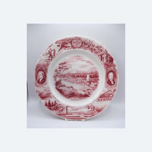 May include: A decorative plate with a red and white design. The plate features illustrations of landscapes, portraits, and text, including "Crater Lake" and "Oregon City". The plate has a scalloped edge.