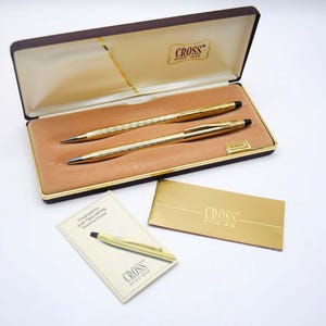 Könnte beinhalten: Ein goldfarbenes Cross Stift- und Bleistiftset in einer Präsentationsbox. Das Set enthält einen Stift, einen Bleistift, eine Garantiekarte und eine Bedienungsanleitung. Die Box ist mit einem beigefarbenen Stoff ausgekleidet und hat das Cross-Logo.