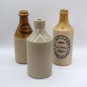 May include: Three antique stoneware bottles in shades of tan and brown. Two bottles have a rounded shape with a narrow neck and a circular label. The third bottle is cylindrical with a wide neck.