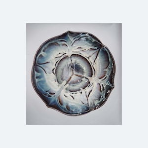 Può includere: Un piatto da portata in ceramica dalla forma organica unica. Il piatto presenta una miscela di tonalità blu, bianche e marroni, con una finitura lucida. Il bordo ha un colore più scuro e contrastante. Questo pezzo decorativo è ideale per servire cibo.