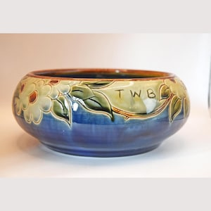 Könnte beinhalten: Eine Keramikschale mit blauem Boden und braunem Rand. Die Schale ist mit floralen und Blattmustern in Grün, Creme und Braun verziert. Die Buchstaben "TWB" sind auf der Seite der Schale zu sehen.